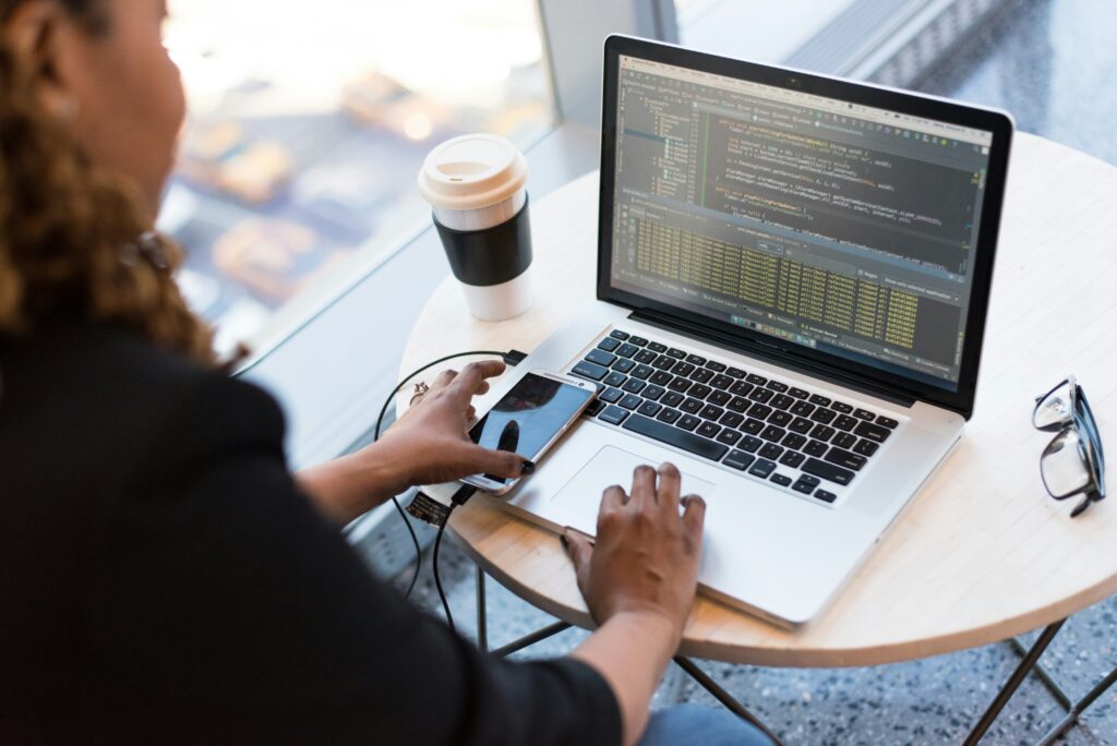 An image of a woman sitting at a table in a cafe. There is software coding on her laptop screen and a mobile phone on the table that she is reaching for as well as a coffee cup to the left of the laptop on the table. It's intended to show a developer working on coding an application as context for the article about the differences between native app development, web app development, and hybrid app development.